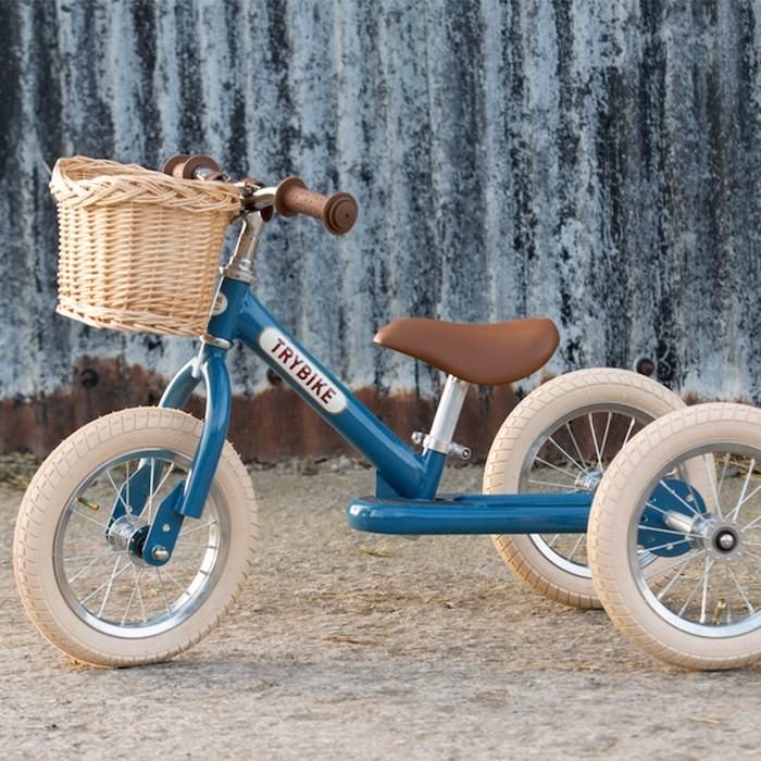 Blue tricycle with brown seat and handles, cream tyres and a beige wicker basket. The background is grey and rusted corrugated iron and a light sandy foreground.