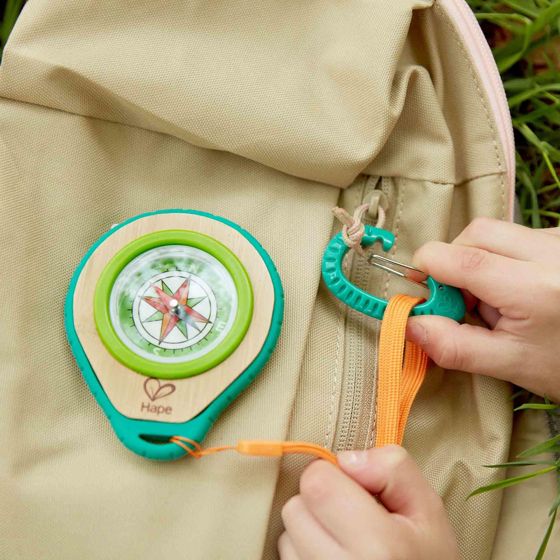 Child's hands attaching the compass using the green carabiner to buff-coloured backpack.