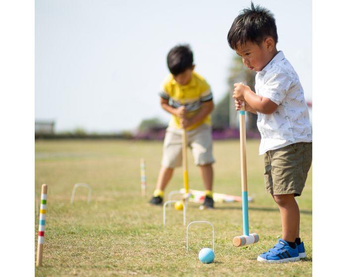 Young boy with dark hair, light top and blue shoes playing croquet with annother young boy in a yellow shirt blurred in the background. The background scene looks like a dusty, not especially green field!