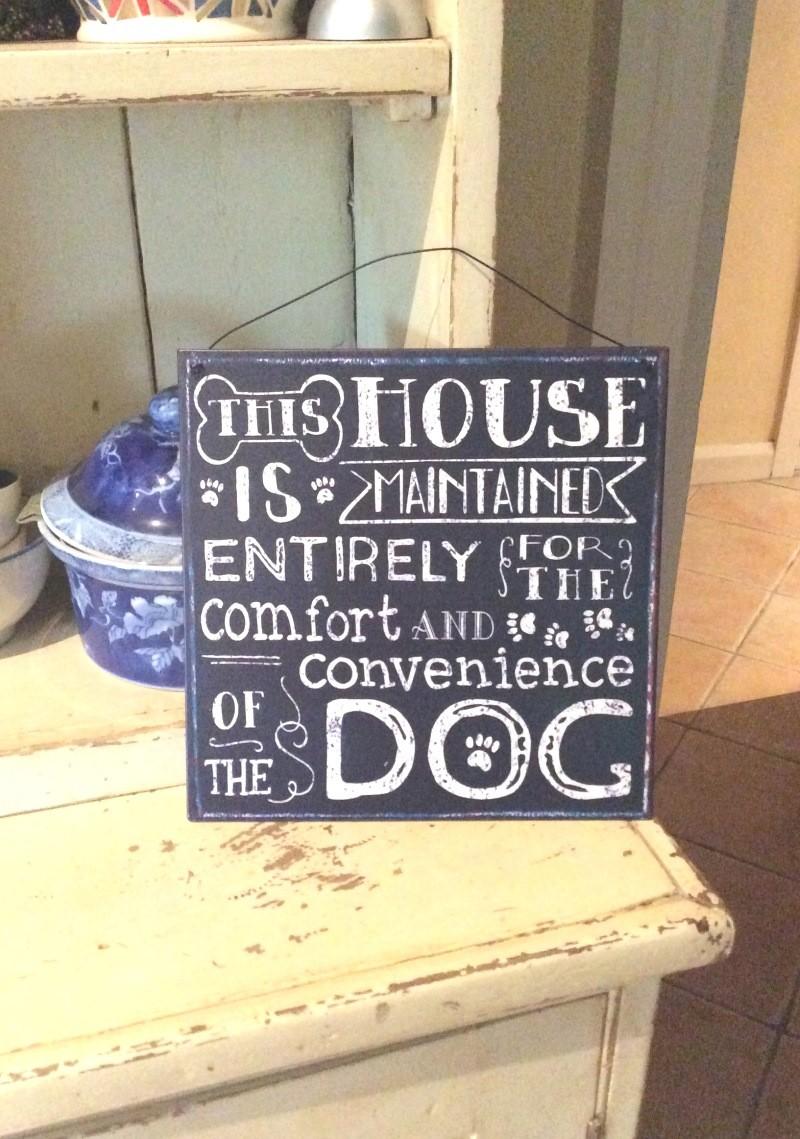 Dark blue metal sign, propped up on a shelf, reads "This house is maintained entirely for the comfort and convenience of the dog."
