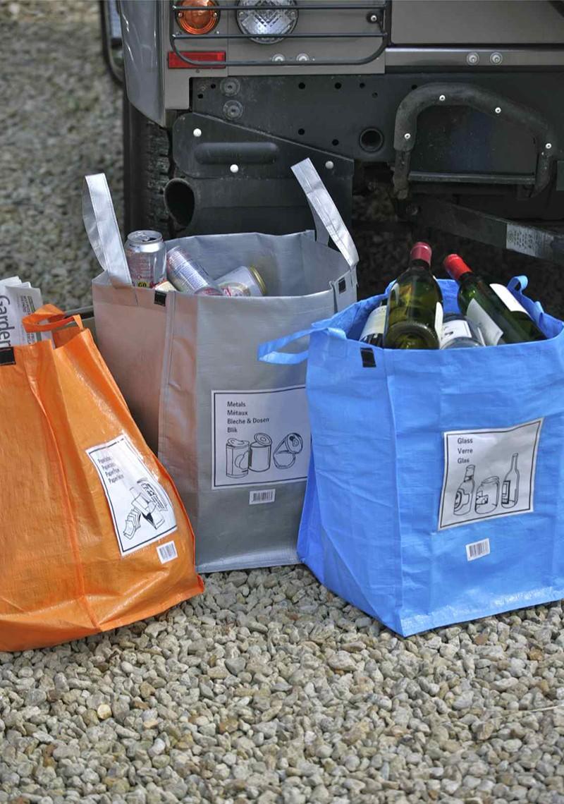 Collection of three recycling bags in orange, silver and blue on a gravel drive behind a vehicle.