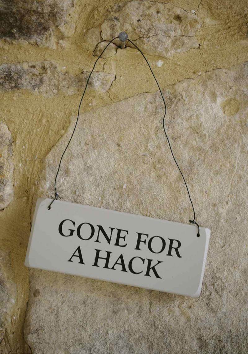 Hand painted, grey/green, rectangular wooden door sign bearing the words "Gone for a hack" hanging on a stone wall.