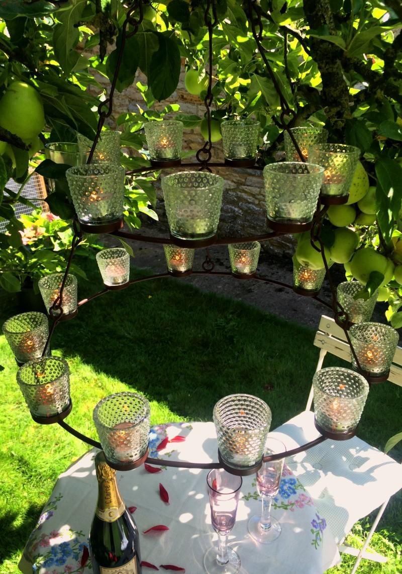Two circles of tealights in bubble glass votives on dark metal ring hanging under a tree above a garden table laid for a party