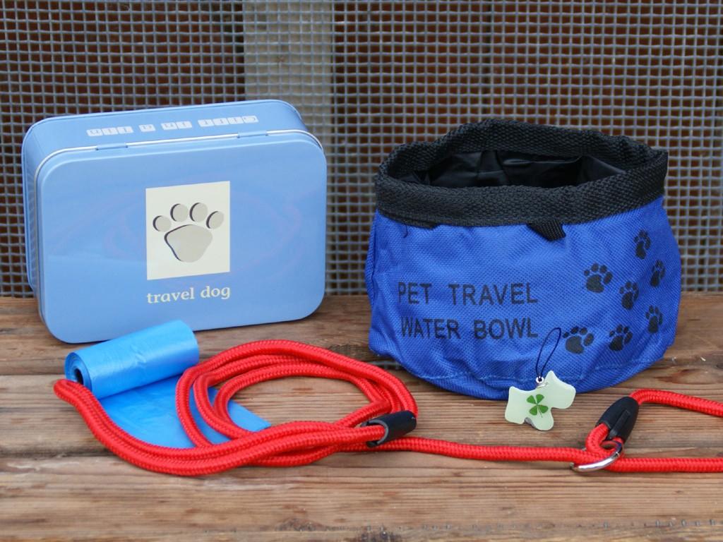 Small Pale blue tin with collapsible water bowl, bright red lead, roll of poop bags and a glow in the dark dog-shaped tag laid out on the table in front.