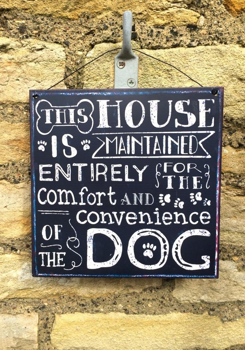 Dark blue metal sign hanging on a stone wall, reads "This house is maintained entirely for the comfort and convenience of the dog."
