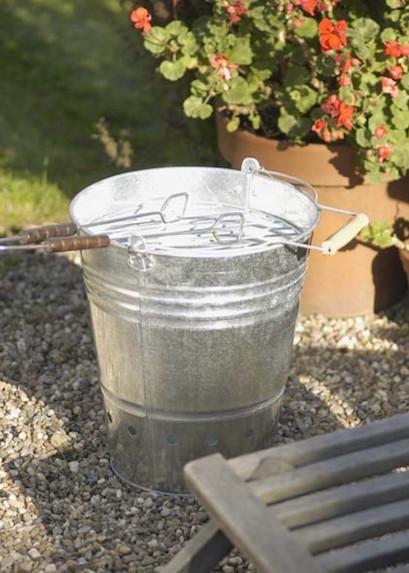 Galvanised metal bucket barbeque with metal wire and wooden handle in a garden setting.