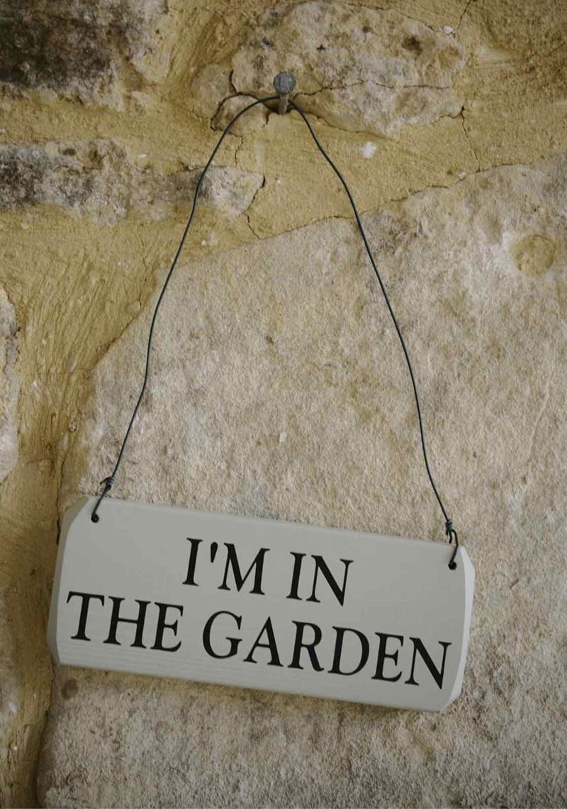 Small painted, wooden sign, with metal wire hanger, bearing the words "i'm in the Garden" hanging on a stone wall.