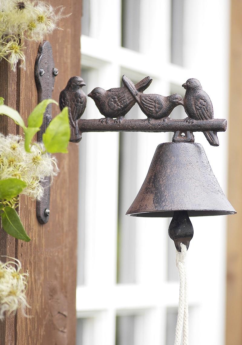 Dark brown cast iron doorbell hanging underneath a pole with a row of little birds sitting on it. Rope bell pull under the bell.