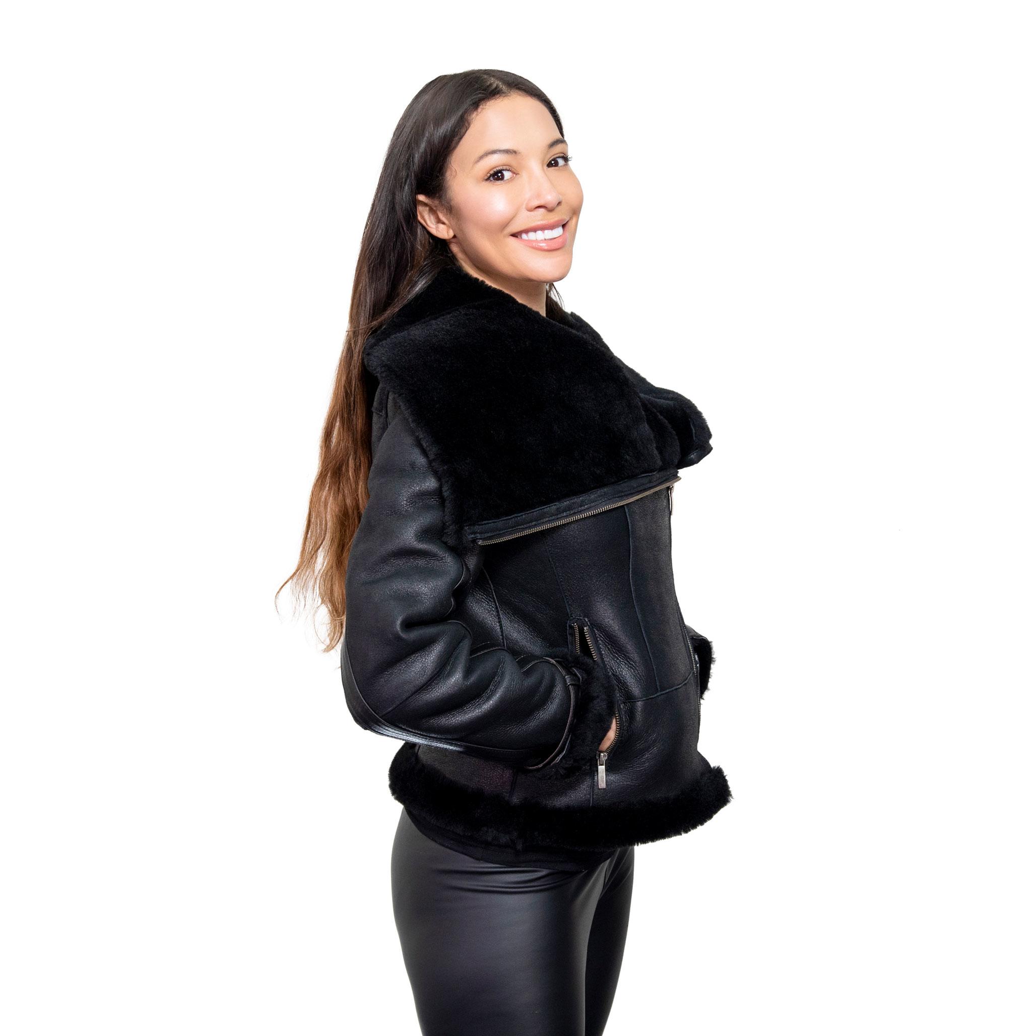 A models stands to the right side, smiling blissfully as she dons a beautiful black sheepskin jacket, with an elegant, large fur lined collar. The outer layer of jacket is in a sleek nappa leather finish, with lush interior sheepskin fur.