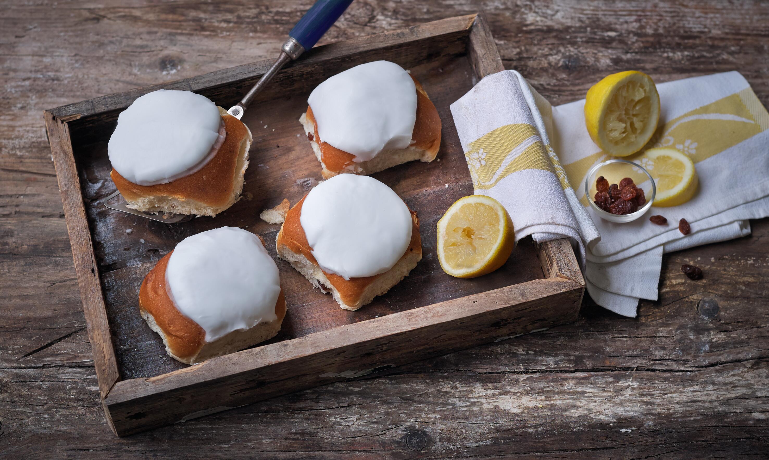 Lifestyle image of four Whitby Lemon Buns with white icing presented on a wooden tray with a spatula holding one bun up. Decorative lemon halves, sultanas and linen included.