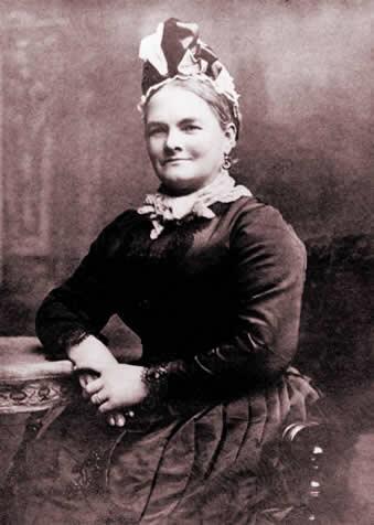 Black and white portrait of Elizabeth Botham, sat on a chair, resting one arm on a table, dressed in Victorian clothes with a headscarf.