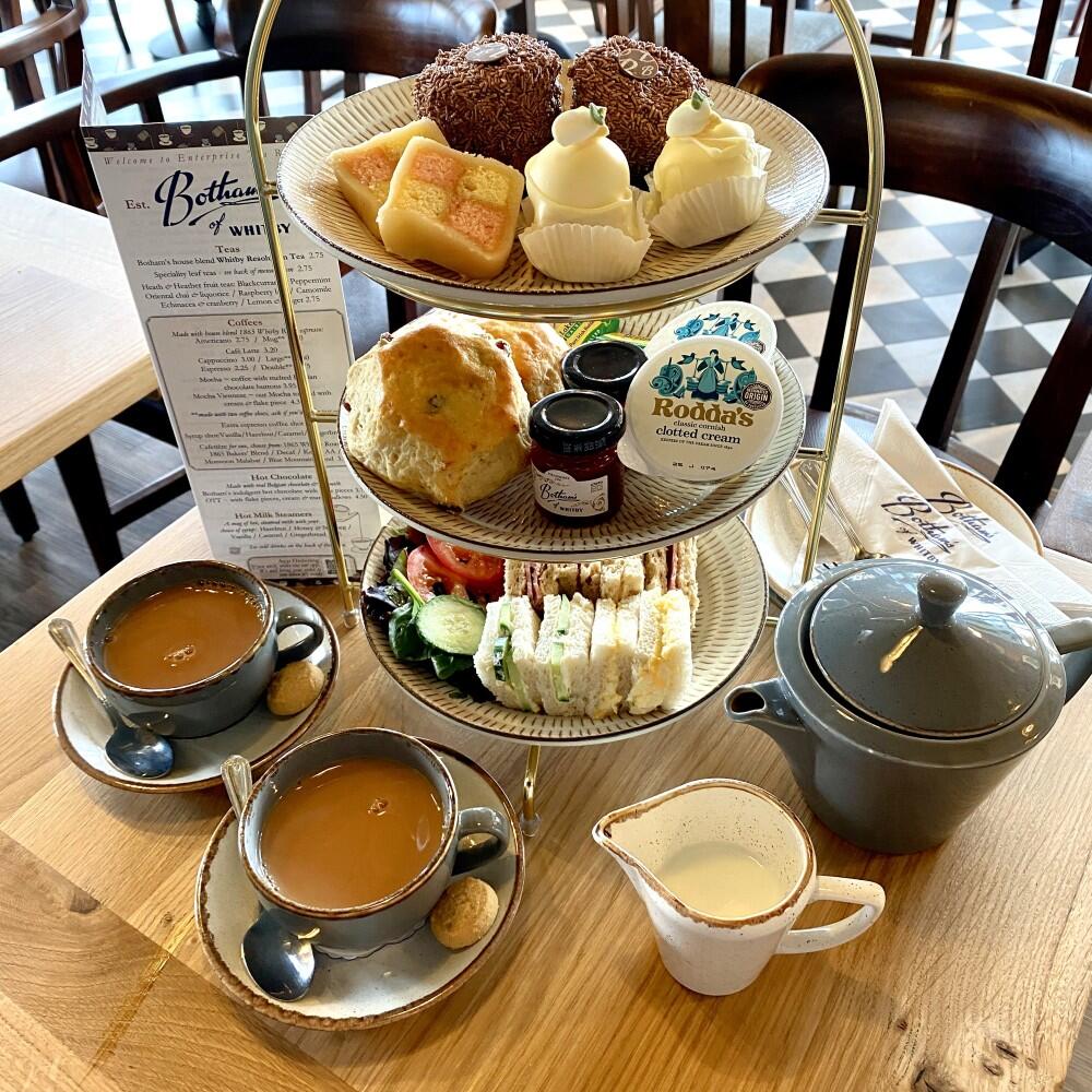Image displays Botham's Afternoon Tea for Two featuring a three tier afternoon tea stand with a selection of mini-fancy cakes on the top tier, a scone, jam & clotted cream middle tier and finally a plate of finger sandwiches and salad garnish on the bottom. Served with house blend Resolution Tea for two, serving plates, cutlery and Botham's branded napkins. A traditional and delicious Afternoon Tea from the Whitby bakers.
