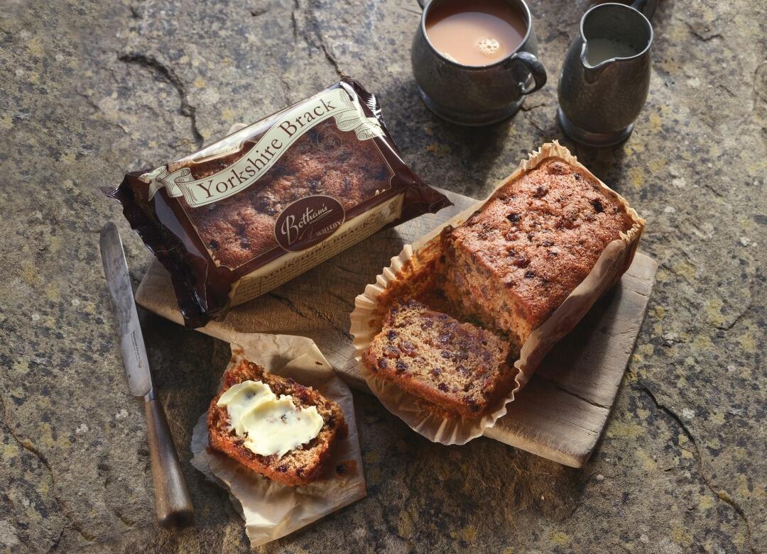 Lifestyle image of Botham's Yorkshire Brack in branded packaging, with one brack resting on a wooden chopping board with a slice cut and generously buttered. Mug of tea adds atmosphere. Yorkshire Brack is a traditional moist tea loaf originating from Yorkshire.