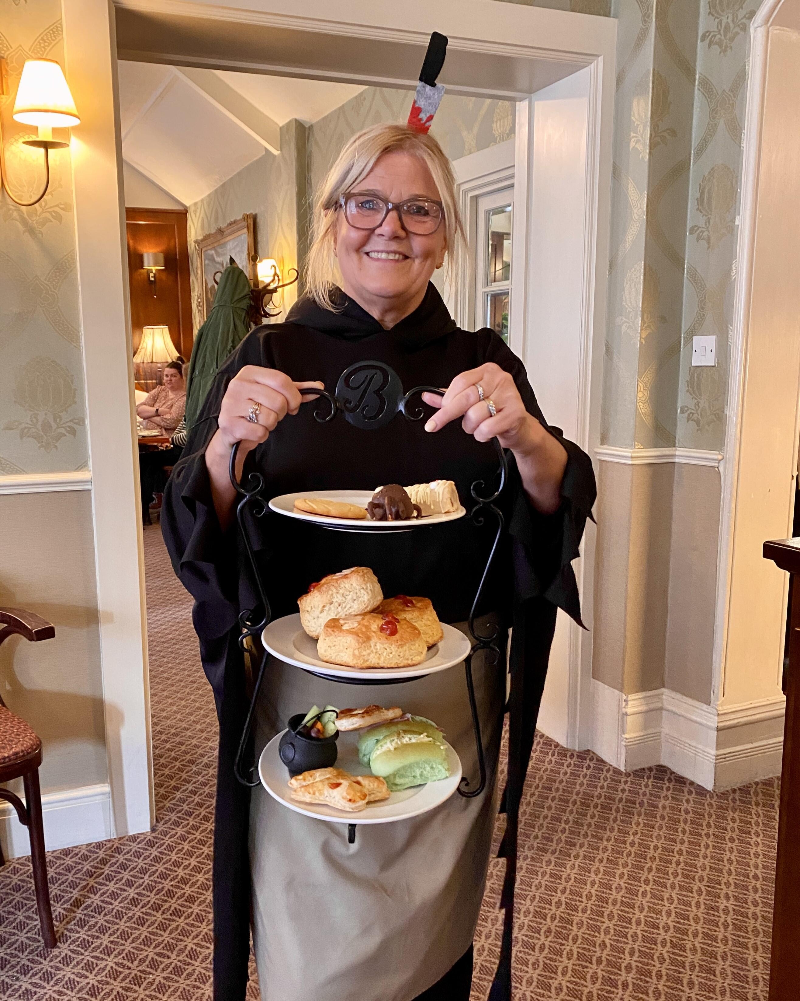 Botham's of Whitby Cafe Assistant in Halloween Fancy dress holding up a three tier cake stand with Botham's Halloween Afternoon Tea presented on it.