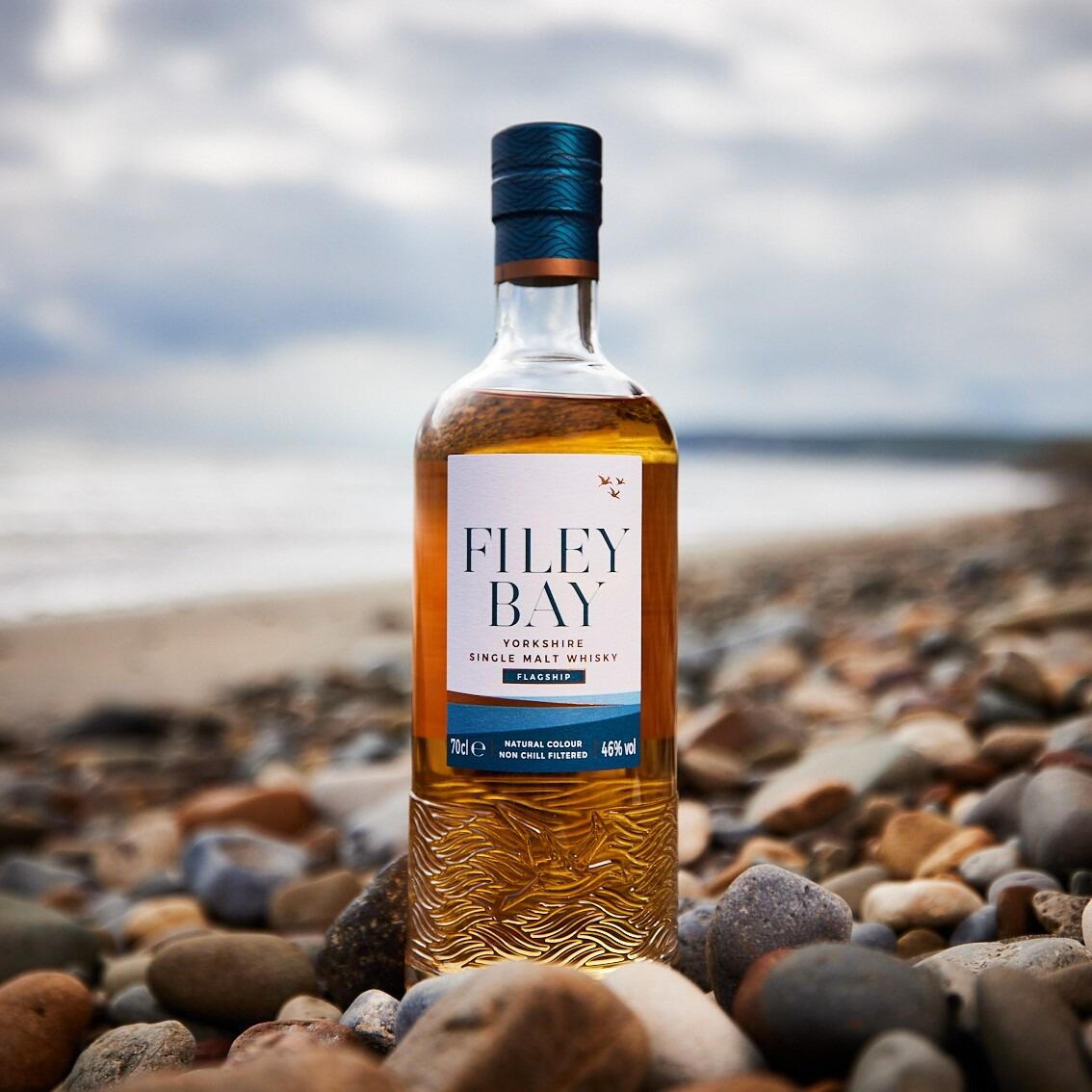 Hero shot of Filey Bay Whisky flagship bottle on the beach with stones in the foreground and the sweeping coastline in the background.