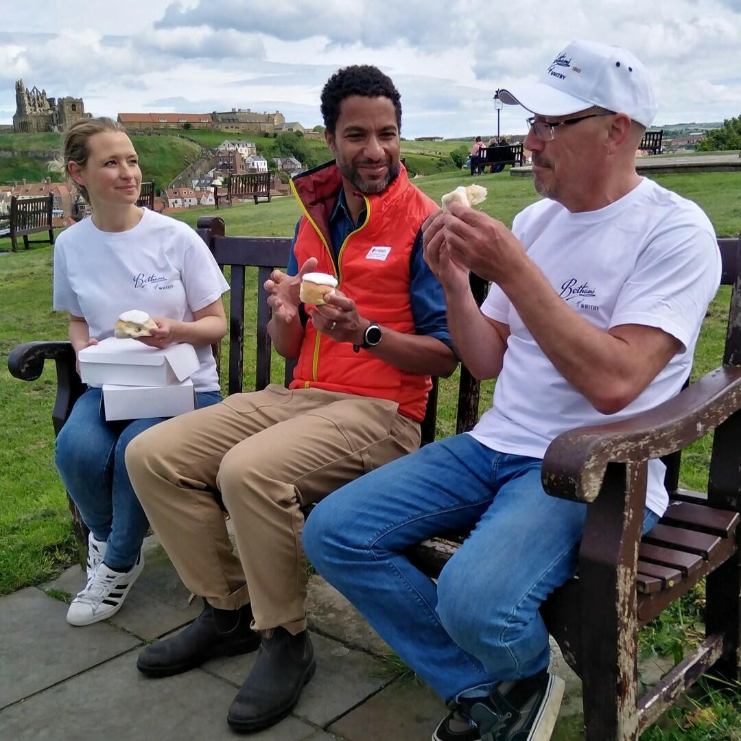 Image of family bakers Jo and Lois sat on a bench on Whitby's West Cliff with Sean Fletcher from BBC One Countryfile, with Jo demonstrating the Whitby way to eat a Whitby Lemon Bun: tearing the bun in half and turning the icing inside to make a sandwich. Whitby Abbey and St Mary's Church stands proud on East Cliff in the background.