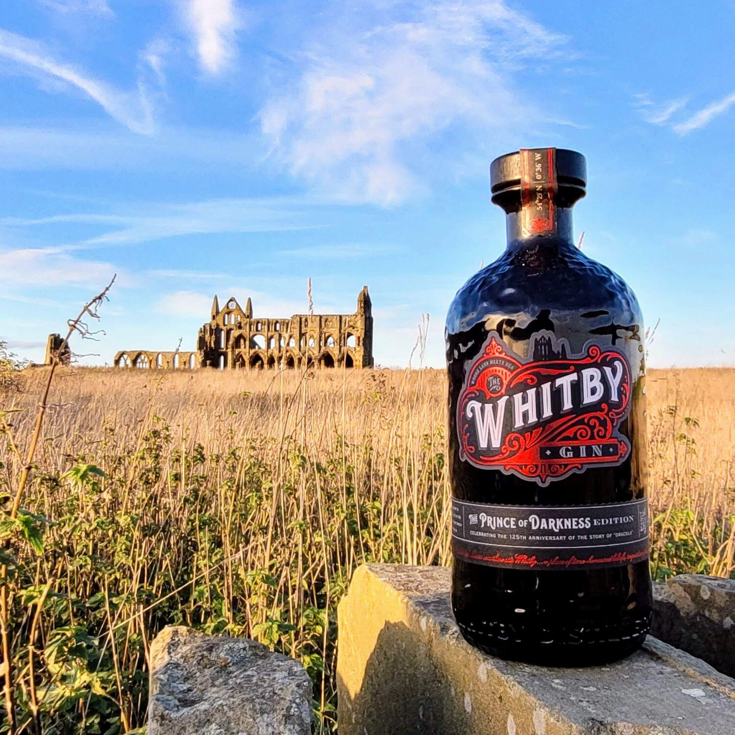 Bottle of The Prince of Darkness Edition Whitby Gin in a black bottle with red label, sat on a wall on Whitby's East Cliff with Whitby Abbey, headlands and blue sky in the background.