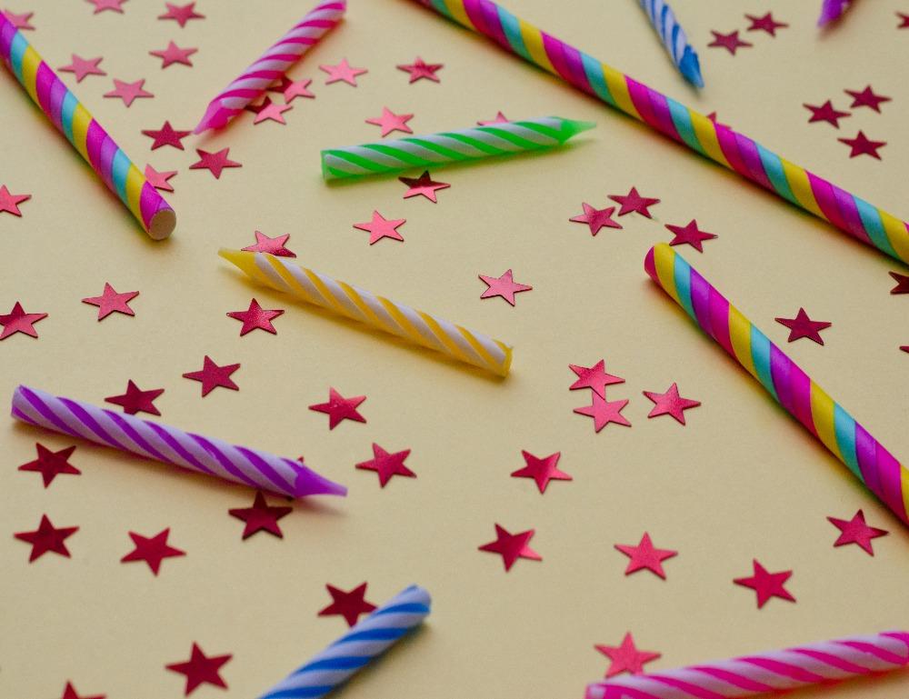 Coloured candles, pink star confetti, striped coloured straws laid on on a yellow background