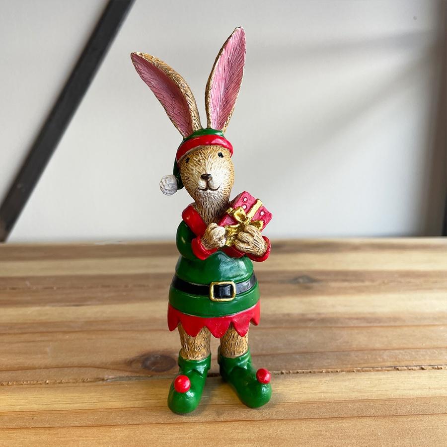 Standing tanned colour rsabbit with big tall ears dressed in a elf suit holding a Christmas present.