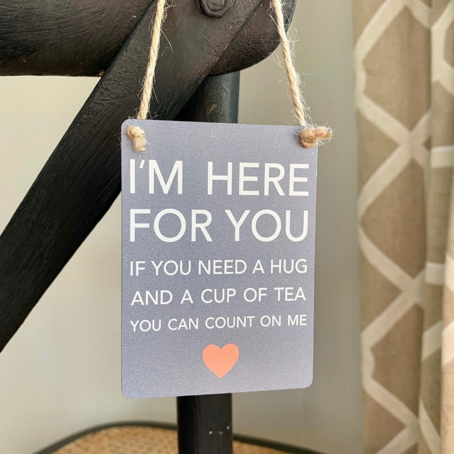 Dark grey small metal sign hanging by jute string. White text reads ' I'M HERE FOR YOU IF YOU NEED A HUG, AND A CUP OF TEA, YOU CAN COUNT ON ME'. Small red heart.