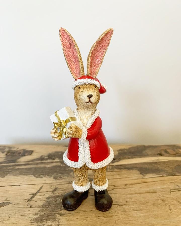 Small standing light brown rabbit, tall pink ears, intricate fur detailing, wearing a Santa coat, Santa outfit with Black boots, holding white gift with gold ribbon around it.