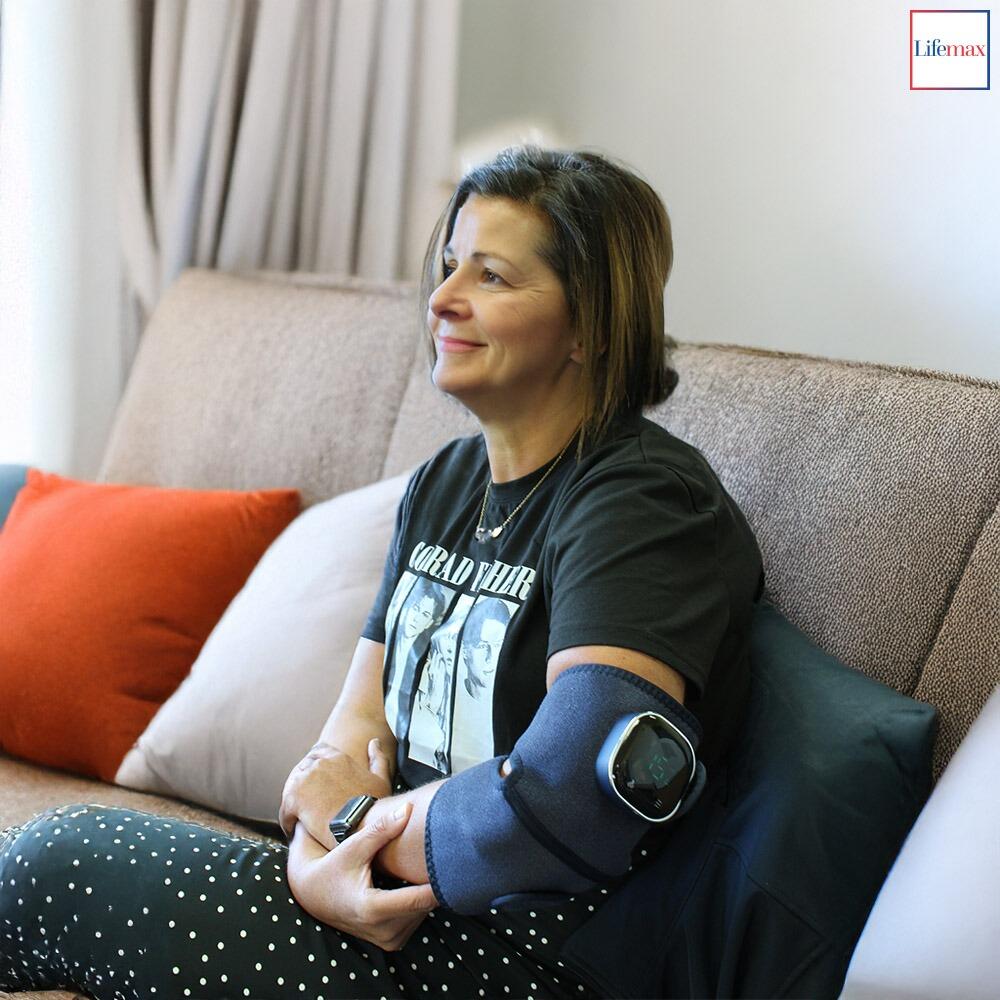 A middle aged lady sat on a sofa using the Lifemax 3 in 1 Heated Vibration Massager on her elbow to treat Arthritis
