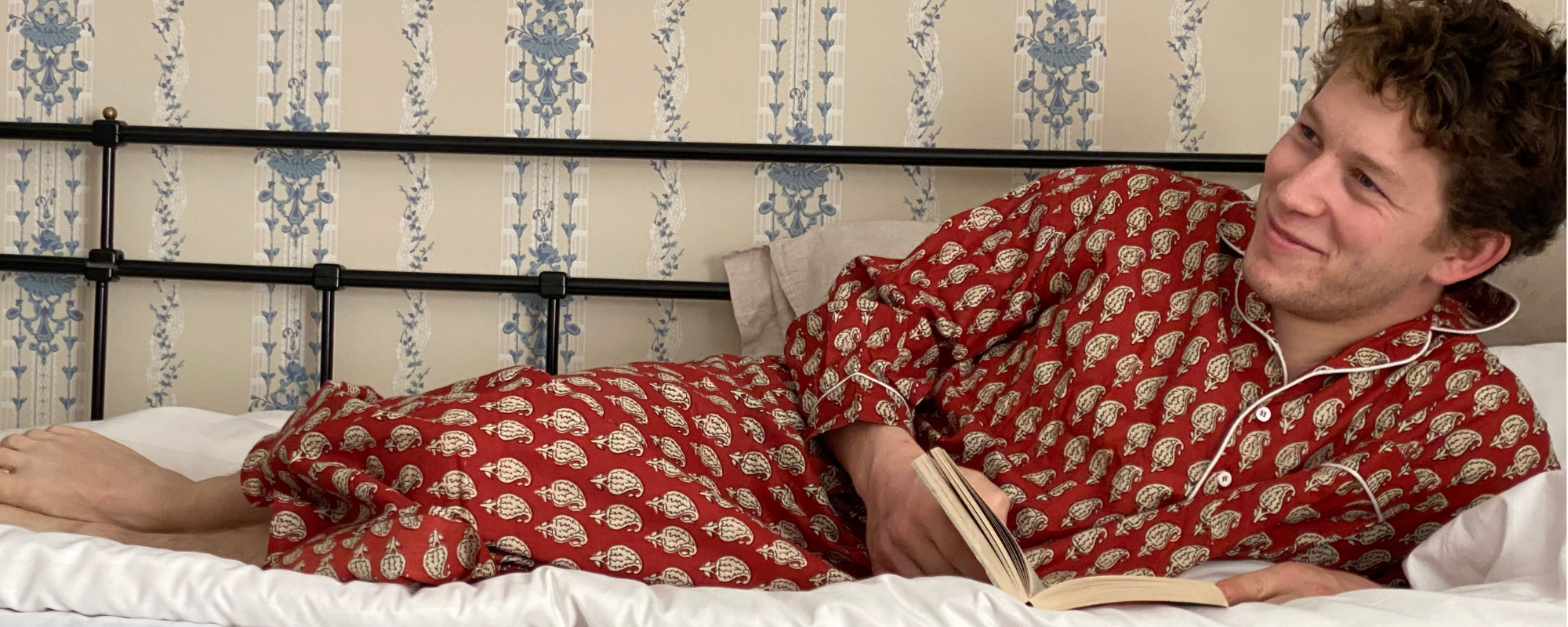 A man wearing a printed cotton nightshirt, lying on a bed, grinning at someone off camera