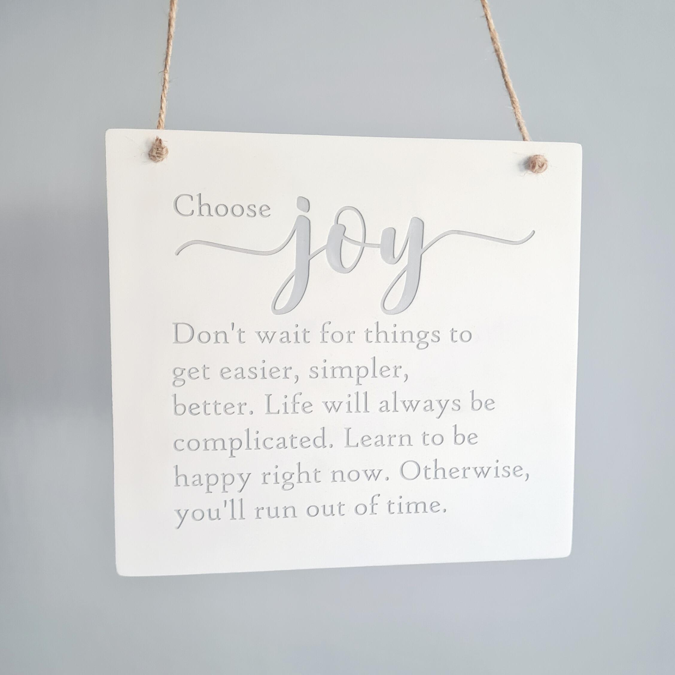 Hanging white clay plaque with the words choose joy, don't wait for things to get easier, simpler, better. Life will always be complicated. Learn to be happy right now. Otherwise, you'll run out of time. The text is imprinted in grey paint with the word joy in large script font. Hanging with jute twine