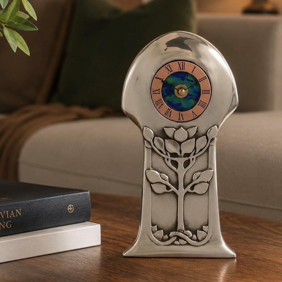 An Arts and Crafts style pewter mantel clock designed by Archibald Knox, featuring a copper dial ring with Roman numerals around a swirling blue and green enamelled centre. The polished pewter case has a rounded mushroom-shaped top and a tapered base decorated with an embossed stylized tree design