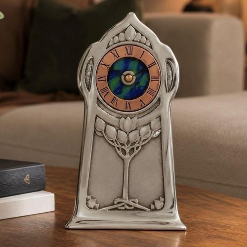 An Arts and Crafts style pewter mantel clock designed by Archibald Knox, featuring a copper chapter ring with Roman numerals surrounding a vibrant blue and green enamelled centre. The polished pewter case has a tall, arched silhouette decorated with an embossed stylized tree design
