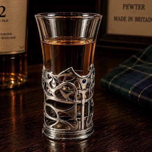 A tall glass shot glass filled with amber-colored liquor, sitting on a dark wooden bar counter. The lower half of the glass is encased in an intricate pewter metal holder featuring an abstract, openwork design of fluid art nouveau swirls and loops. Small, colourful enamel dots in shades of blue and green are subtly inset as accents within the pewter framework