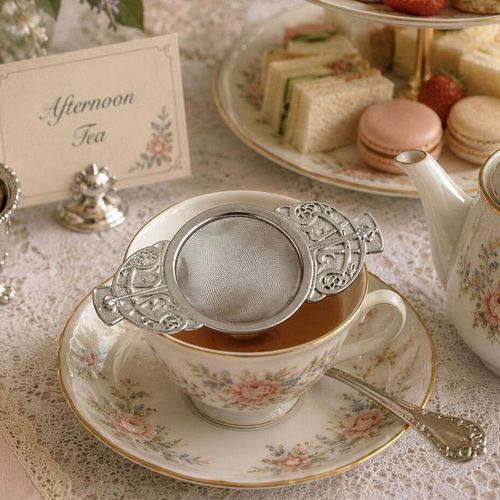 A stainless steel mesh tea strainer with ornate pewter handles featuring an Art Nouveau Celtic knot design, resting across a floral porcelain teacup during an afternoon tea service