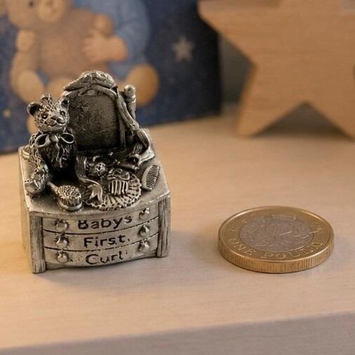A small, detailed pewter keepsake box shaped like a miniature dressing table or chest of drawers, shown next to a British one pound coin for scale on a light wooden shelf. The front of the drawers is engraved with the words "Babys First Curl". Sitting on top of the counter is a sculpted teddy bear figure with a bow, a small vanity mirror, a comb, and a hairbrush