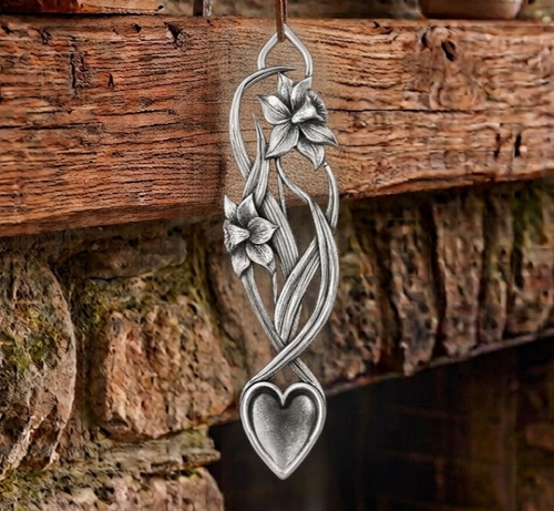 Handcrafted pewter Welsh lovespoon hanging against a wooden beam and stone fireplace, featuring an openwork design of two blooming daffodils with flowing stems and long leaves, leading down to a heart-shaped spoon bowl.