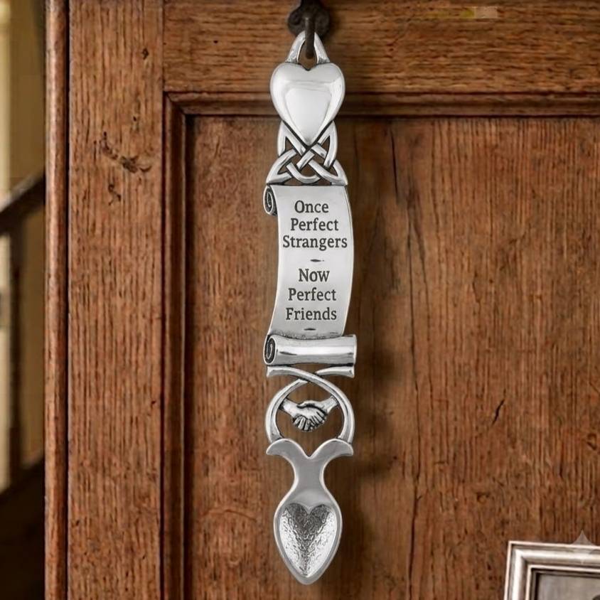 Handcrafted pewter Welsh lovespoon hanging on a wrought iron hook against a wooden panelled wall, featuring a polished top heart, Celtic knotwork, an unrolled scroll engraved with "Once Perfect Strangers Now Perfect Friends", handshaking friendship hands motif, and a textured spoon bowl.