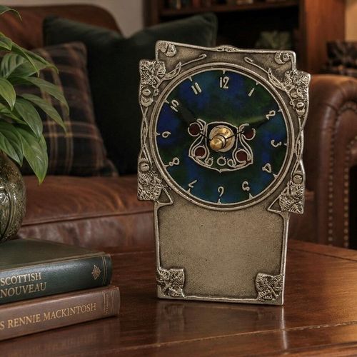 An Arts and Crafts style pewter mantel clock designed by Archibald Knox, featuring a vibrant blue and green enamelled circular face with stylized numerals and a Celtic-inspired centre motif. The rectangular pewter frame is decorated with embossed leafy corner details