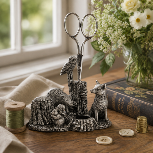 Woodland-themed pewter sewing station with integrated scissors holder, pin cushion holder, and thimble holder, displayed on a sewing table beside thread, buttons, and wildflowers.