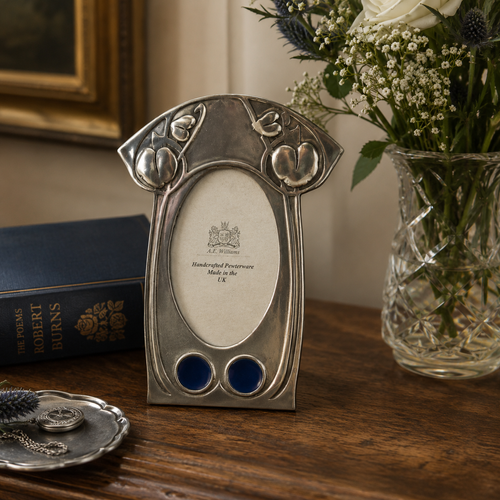 Archibald Knox-inspired pewter picture frame displayed on a polished wooden table beside fresh flowers, a vintage book, and decorative silver accessories in an elegant heritage-style interior.