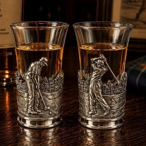 A tall glass shot glass filled with amber-colored liquor, standing side-by-side on a dark wooden bar counter. The lower half of each glass is encased in a detailed pewter metal holder featuring a relief-sculpted golfing scene against a background of trees and fairways. The glass on the left shows a golfer in a cap bending over to line up a putt with his golf club, while the glass on the right depicts a golfer mid-swing just after hitting the ball, with a golf flag visible on the green behind him