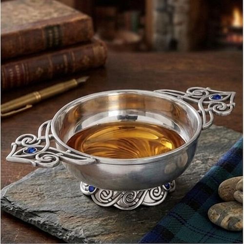 Traditional pewter quaich bowl filled with whiskey, featuring ornate Celtic knotwork handles inlaid with blue gemstones, displayed on a slate mat with a tartan cloth and vintage books