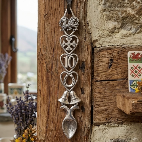 Handcrafted pewter 10th anniversary Welsh lovespoon hanging on a rustic wooden beam, featuring intricate carved symbols including a Welsh dragon, Celtic cross, the number 10, a horseshoe, wedding bells, and a heart-shaped spoon bowl