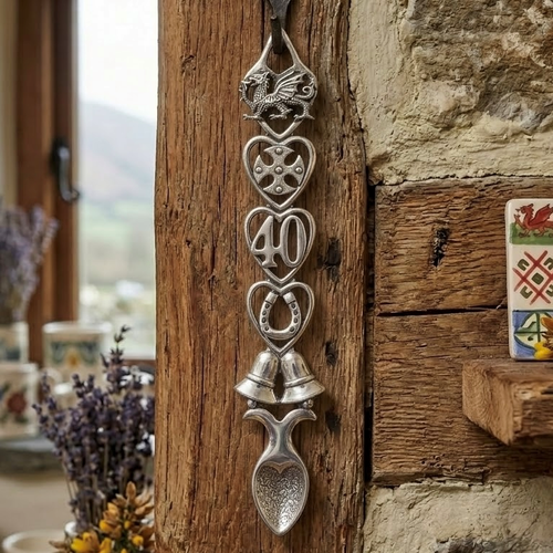 Handcrafted pewter 40th anniversary Welsh lovespoon hanging on a rustic wooden beam, featuring detailed symbols including a Welsh dragon, Celtic cross, the number 40, a horseshoe, wedding bells, and a heart-shaped spoon bowl.