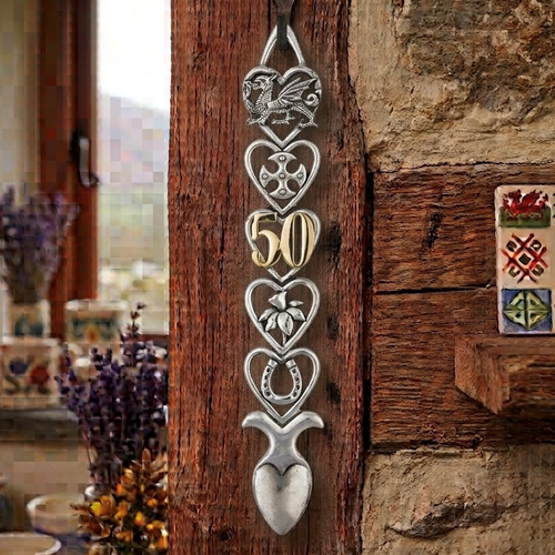 Handcrafted pewter 50th birthday Welsh lovespoon hanging on a rustic wooden beam, featuring detailed symbols including a Welsh dragon, Celtic cross, a gold-toned number 50, a daffodil, a horseshoe, and a heart-shaped spoon bowl.