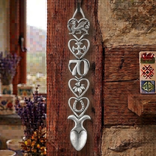 Handcrafted pewter 70th birthday Welsh lovespoon hanging on a rustic wooden beam, featuring detailed symbols including a Welsh dragon, Celtic cross, the number 70, a daffodil, a horseshoe, and a heart-shaped spoon bowl.