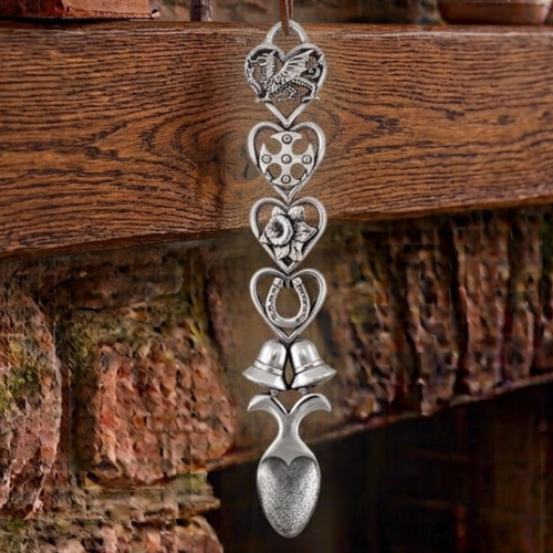 Handcrafted pewter Welsh lovespoon hanging against a wooden beam and stone fireplace, featuring heart-shaped frames enclosing a Welsh dragon, a Celtic cross, a daffodil, a lucky horseshoe, and twin wedding bells above a textured spoon bowl.