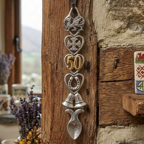 Handcrafted pewter Welsh lovespoon hanging on a rustic wooden beam beside a stone wall, featuring heart-shaped frames enclosing a Welsh dragon, a Celtic cross, gold-plated number 50 digits, a horseshoe, and twin wedding bells above a textured spoon bowl.