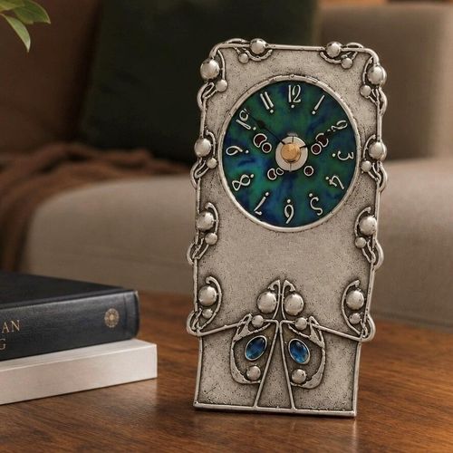 An Arts and Crafts style pewter mantel clock designed by Archibald Knox, featuring a circular face with a swirling green and blue enamelled background and stylized white numerals. The rectangular pewter case is embellished with raised, organic line work and beaded borders, accented by two bright blue enamelled drops near the base