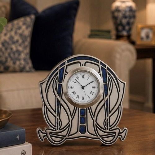 An Arts and Crafts style pewter mantel clock designed by Archibald Knox, featuring a rounded, symmetrical silhouette with sweeping Art Nouveau line work. A white clock face with black Roman numerals is centred within the frame, accented by inset bands and vertical segments of deep blue enamel