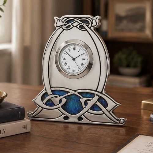 An Arts and Crafts style pewter mantel clock designed by Archibald Knox, featuring a bulbous, horseshoe-shaped frame with an integrated wide base. The clock has a circular white face with black Roman numerals and is accented with intricate Celtic knotwork, including an enamelled top detail and a large, vibrant blue and green enamelled panel stretching across the bottom