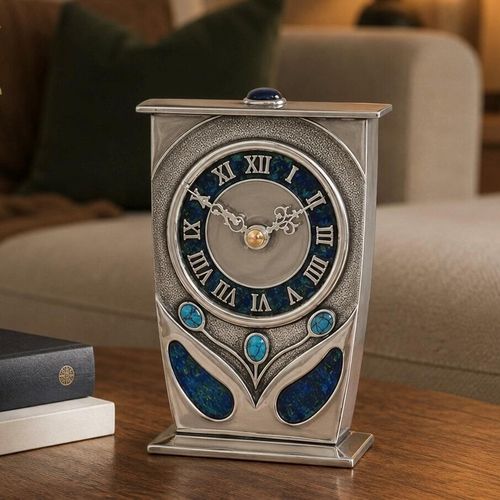 An Arts and Crafts style pewter mantel clock designed by Archibald Knox, featuring a tapered rectangular frame with a flat top adorned with a blue cabochon gem. The large circular clock face has a blue and green enamelled chapter ring with silver Roman numerals, while the base is embellished with three turquoise cabochons and two teardrop-shaped panels of blue enamel inlay, resting on a wooden table surface